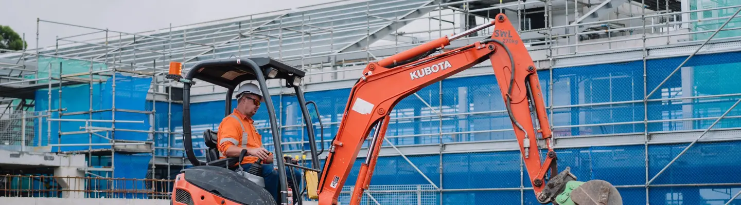 Man operating an excavator to excavate land near safety barriers on an active construction site.