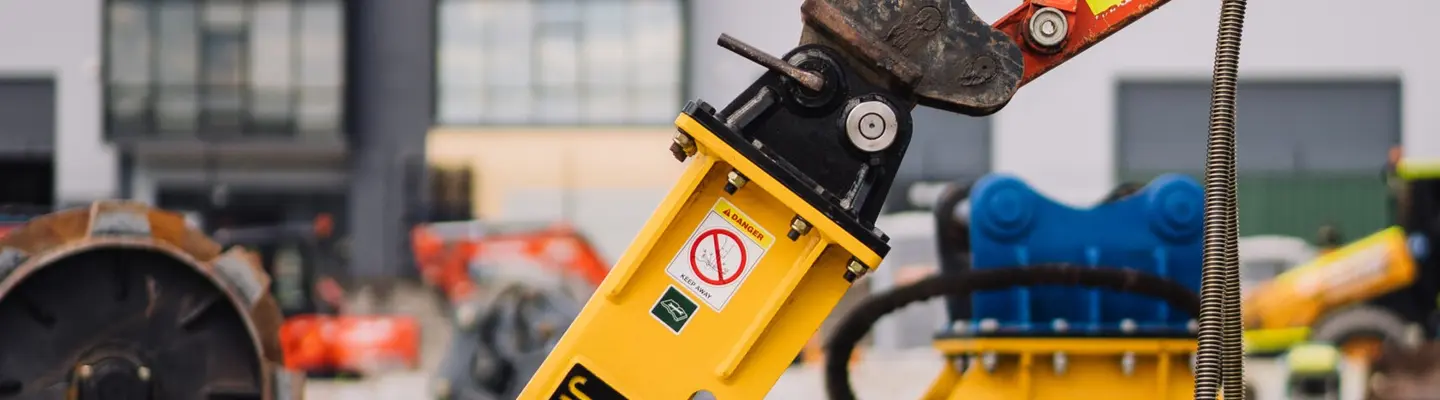 Excavator with a drill attachment at the tip, used for breaking ground at a construction site with safety measures in place.