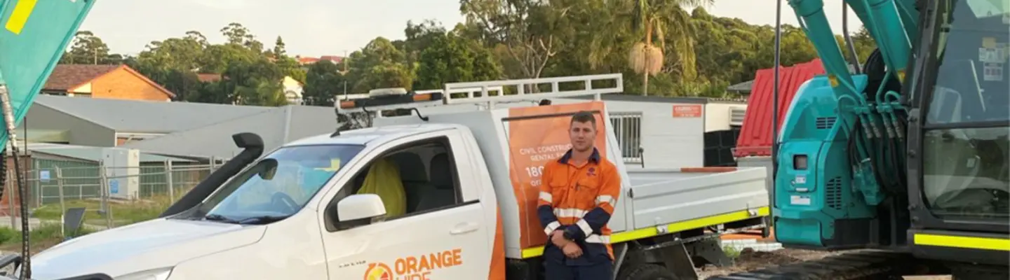 Man standing beside an excavator and Orange Hire vehicle at a construction site with safety barriers in place.