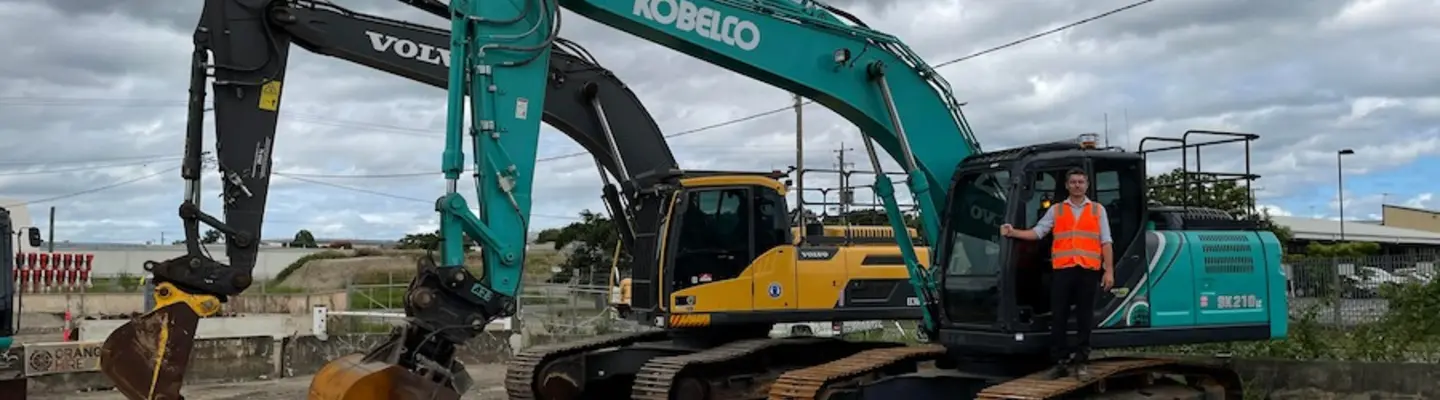 Man standing in a parked excavator at a construction site with safety barriers in the background.