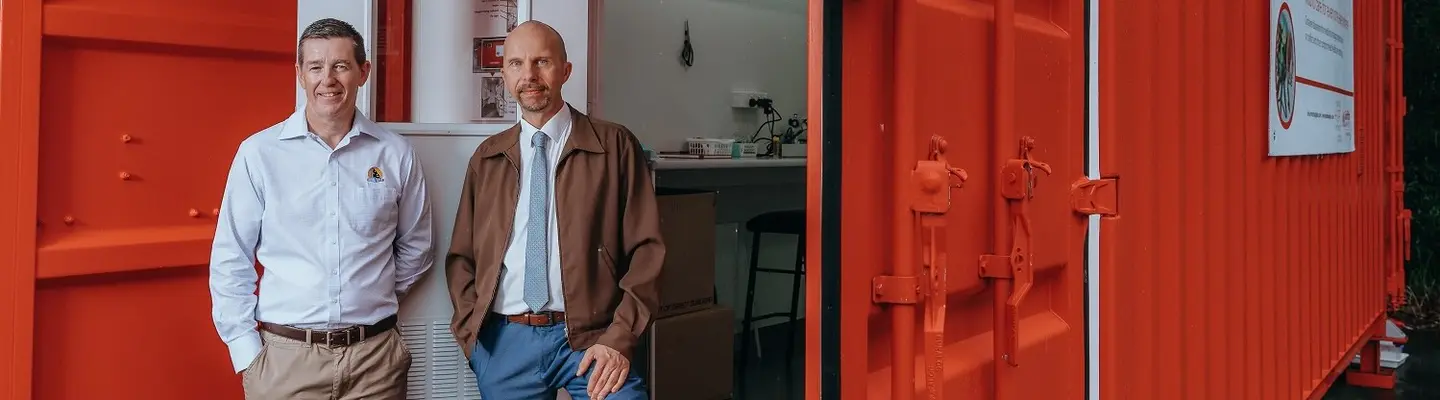 Two men standing beside an open red container office with a visible interior workspace.