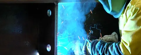 A welder in protective gear creating a bright blue arc while fabricating a large metal beam.