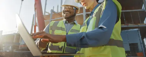 Two workers on a construction site using a laptop.