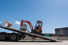 Man driving construction equipment loaded on a trailer truck at a worksite with safety barriers in the background.